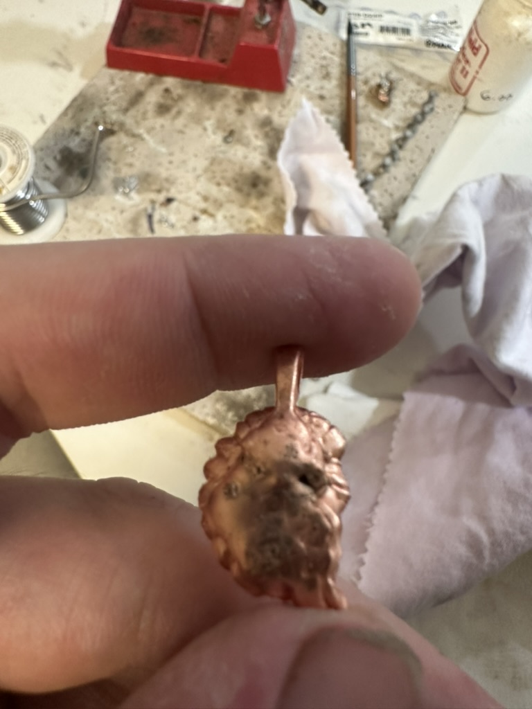 Hand holding a small copper piece with solder tools and work surface in the background