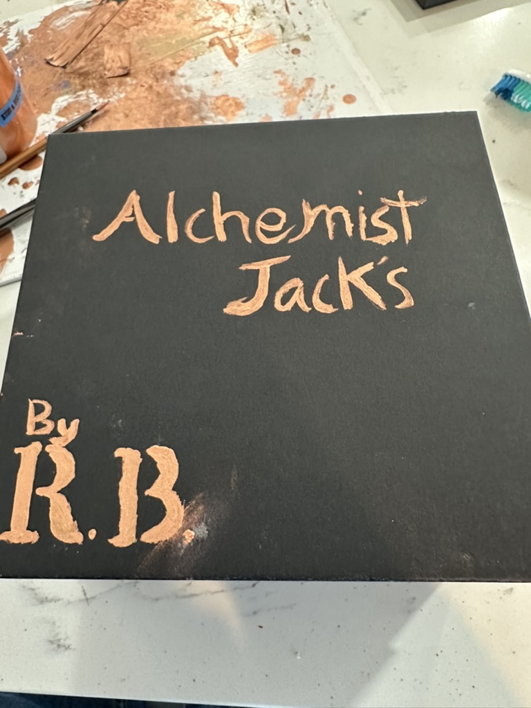 Black square with hand-painted metallic copper lettering reading Alchemist Jack’s and By R.B.